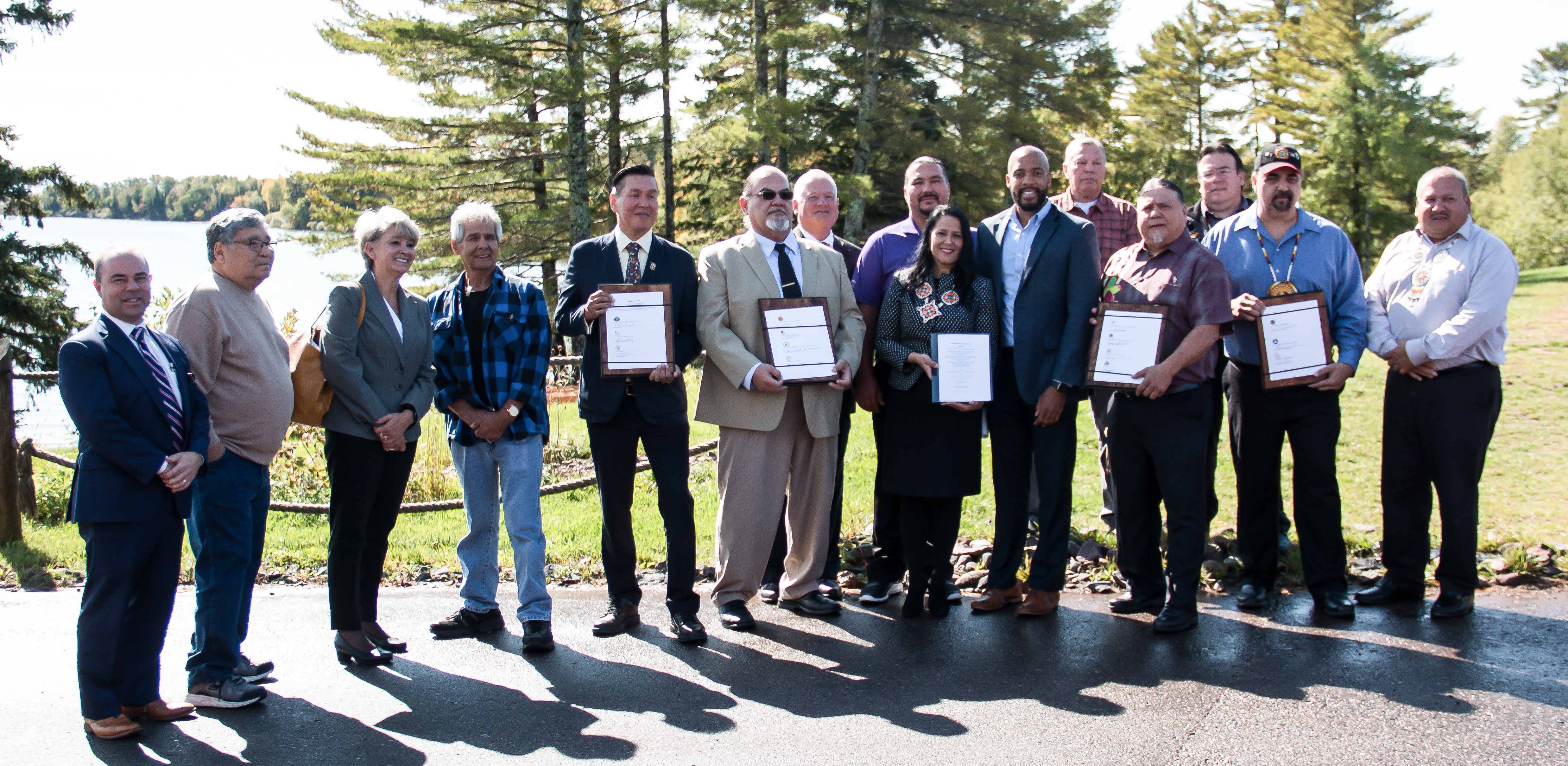 Representatives of tribal governments and the lt. governor pose for a photo in front of trees and a lake.